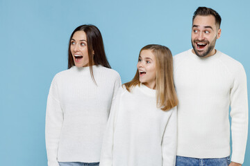 Surprised young parents mom dad with child kid daughter teen girl in white sweaters keep mouth open looking aside isolated on blue background studio portrait. Family day parenthood childhood concept.