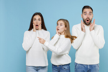 Amazed young parents mom dad with child kid daughter teen girl in white sweaters point index finger aside put hand on cheeks isolated on blue background studio portrait. Family day parenthood concept.