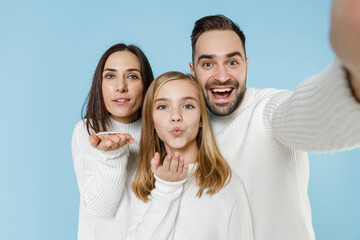 Close up of pretty young parents mom dad child kid daughter teen girl in sweaters doing selfie shot on mobile phone blowing sending air kiss isolated on blue background. Family day parenthood concept.