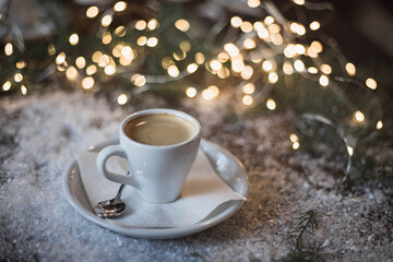 A cup of espresso surrounded by New Year's lights and pine branches in the snow. Dark background. Christmas mood