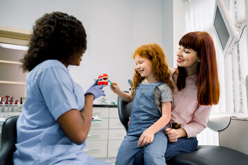 Female African doctor demonstrating proper dental care to young mother and schoolgirl child. Happy kid cleaning teeth with toothbrush and having fun at modern dentist's office