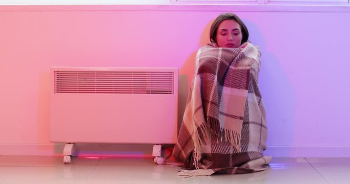 Young woman wrapped in warm plaid sitting near switched on electric heater in room