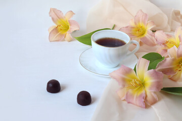 A cup of coffee with two chocolates, daylily flowers on a delicate scarf on a white background, a place for text, a side view-the concept of pleasant moments of relaxation over a cup of coffee