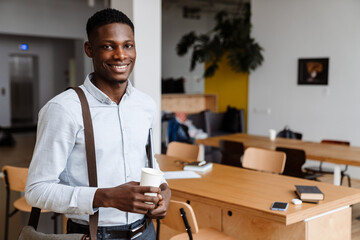 Afro american pleased man drinking coffee while standing with laptop
