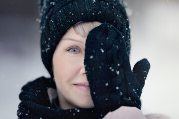 A young beautiful girl covers half her face with her hand in a mitten, playing with snow, catches snowflakes