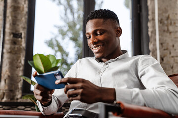 Afro american young man smiling and holding smartphone in armchair