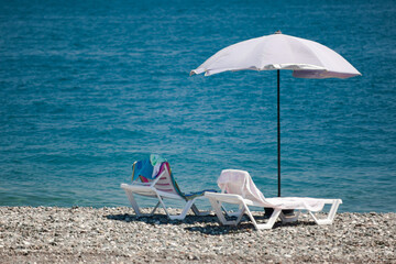 Beach umbrella from the sun and two sun loungers stand on a glyses beach against the blue sea, on deckchairs are towels