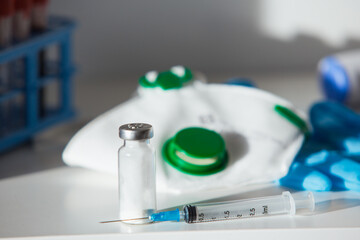 Close up of syringe with Covid-19 vaccine on a white table with protective mask and gloves and blood tube rack.