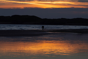 Silhouette of dog walkers on a beach at sunset with hills in the background. Taken at Beaumaris, Anglesey, Wales, with a view across the Menai Straits to the edge of Snowdonia national park.