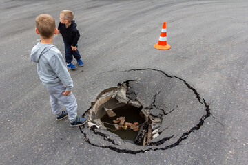 Children play on road near huge deep sinkhole in asphalt surface. Risk of injury to child. Damaged sink hole on city street
