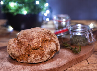 Fototapeta na wymiar Home baked loaf of bread on wooden board, rosemary branch, glass of home-grown spices, Christmas lights soft focus background