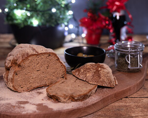 Two slices home baked loaf of bread, wooden board, rosemary branch, black bowl, green olives, glass of home-grown spices, Christmas lights, wine bottle with red ribbon and bow, soft focus background