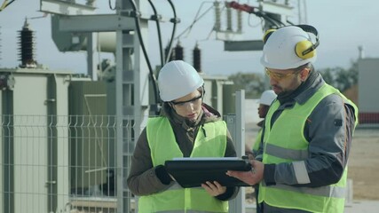 A man and a woman energy engineer next to a transformer discuss work issues, while two workers are talking in the background.