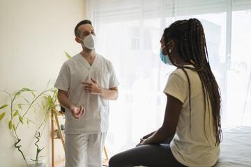 Physiotherapist with mask talking to an african patient sitting on a stretcher