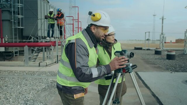 A man and a woman power builders next to a geodetic device, a total station, discuss work issues, while in the background two engineers are talking next to a powerful transformer.