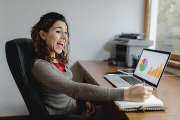 Close-up young caucasian business woman portrait working at office desk. Smiling lady taking notes on notebook paper while talking about new strategy plan studying graphs on desktop computer.
