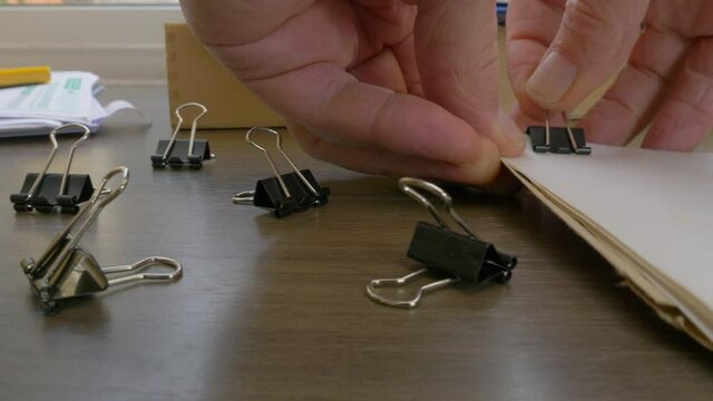 Closeup POV dolly shot past sprung metal clips (bulldog clips / springback clips / binder clips), to a man&rsquo;s hands using one to clamp some documents together.