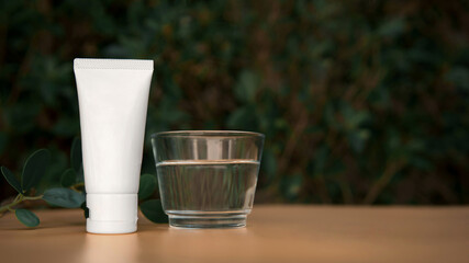 Facial Skincare white tube product with blank label next to transparent glass of pure water with blurred bush nature background and copy space