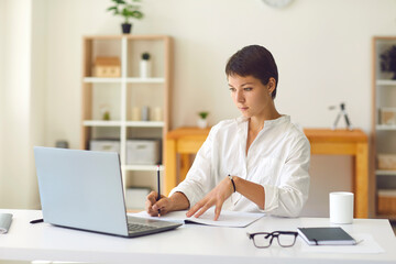 Young woman looking at laptop and making notes during online lesson