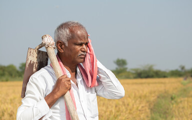 Indian farmer with hand hoe on his shoulder holding towel to his face due to high temperature while working on agriculture land. © WESTOCK