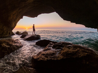 
Man from behind against the light inside a cave observes the sunset overlooking the sea, warm and...