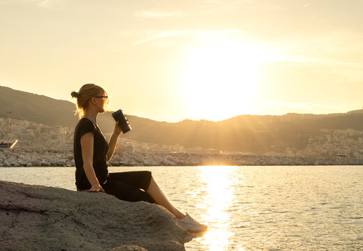 Woman with tumbler cup sitting on a rock of cliff with sea and cityscape on background during sunset, copy space © 22Imagesstudio