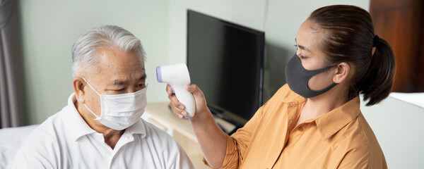 Middle age woman thermal scanning her senior man for fever, COVID-19, coronavirus infection, concept of home health care, family new normal social distancing while awaiting for vaccine
