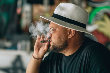 Profile photo. Close up portrait. A bearded man in a black and white panama smoking a cigar and cigarette smoke flying off next to his face.