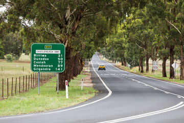 A car travels on a long straight country road through an avenue of trees on a single lane section of the Castelreagh Highway in rural New South Wales, Australia.