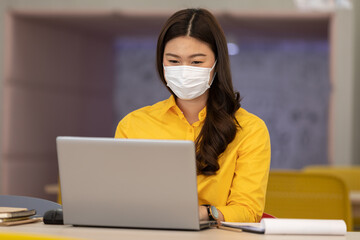 New normal of Asian woman in yellow shirt wearing surgical face mask working with computer laptop thinking to get ideas and requirement in Business startup at modern office or Co-working space