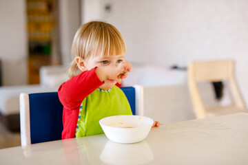 Cute baby 1,4 years old sitting on high children chair and eating vegetable alone in white kitchen