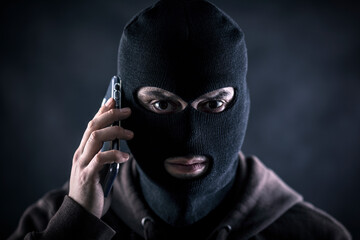 Criminal wearing black balaclava and hoodie with a smartphone in the dark