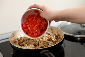 young housewife fry meat with tomatoes in a pan and electric stove. Cooking dinner at home.