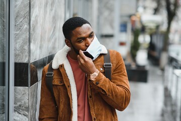 Close-up portrait of a stylish African American guy stands alone in the street in a protective mask on his face. Precautionary measures