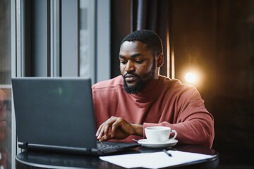 Portrait of thoughtful and concentrated young African American copy-writer, working on new articles using his laptop, and, looking at papers on a cafe table