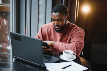 Portrait of thoughtful and concentrated young African American copy-writer, working on new articles using his laptop, and, looking at papers on a cafe table