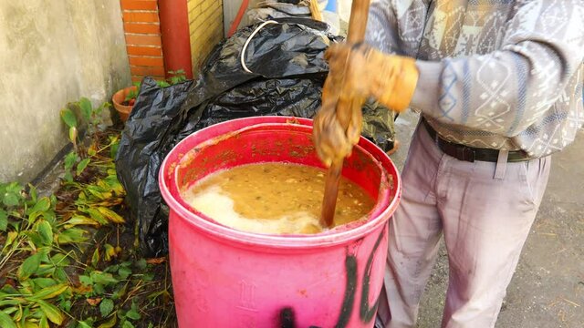 Man is mixing fruit marc made of chopped apples in plastic barrel using hoe. Shuffle process is for better fermenting alcohol, making alcoholic beverages such as brandy.