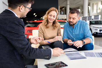Mid-adult couple talking with sales agent and signing contract for new car in modern car showroom.