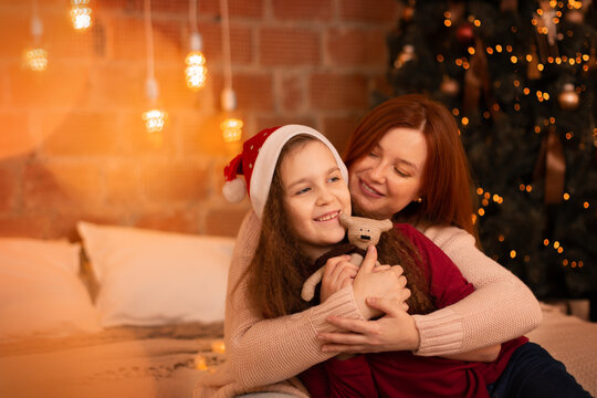 Happy young mother sitting on bed near decorated Christmas tree and hugs her 8 years daughter