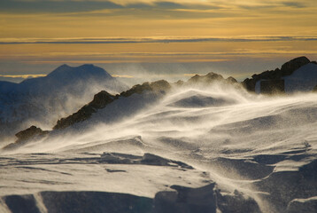 Sommet d'une montagne déserte où le vent souffle, balayant la neige fraiche qui se retrouve en suspension. On peut ressentir le froid. Aucun randonneur ou skieur à l'horizon.
