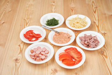 Seven white plates with chopped pizza ingredients on a light wooden background. Close-up. View from above.