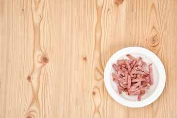 On a light wooden tabletop there is a white plate with sausage cut into strips. Copy space.