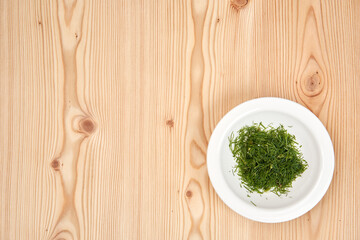 On a light wooden tabletop a white plate with chopped dill. Copy space.
