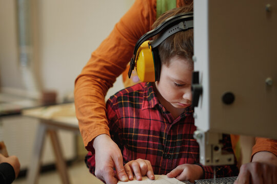 cute caucasian boy in headphones carving a wooden christmas tree with a band-saw in joinery. Woman teacher helping him. Image with selective focus