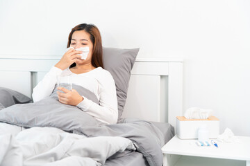 Young Asian Woman with seasonal infections Cold Blowing Her Nose and sneezing into Tissue with headache lying under the blanket in bed with high fever and a flu.