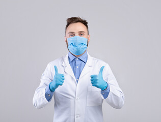 Millennial doctor in uniform, face mask and gloves looking at camera and showing thumbs up gesture over grey background