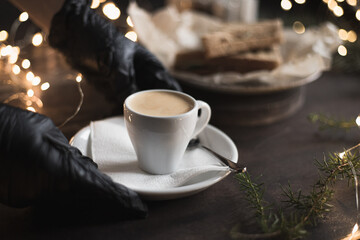 A waiter in medical gloves puts a cup of espresso on the table in coffee shop, Christmas bokeh lights. Selective focus