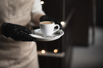 Barista wearing black medical gloves carries a cup of espresso in a coffee shop. Selective focus. Protecting health