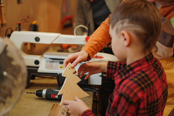 side view of cute caucasian preschooler boy holding a wooden christmas tree with a hole he  made himself using electric drill. arts and crafts class in joinery Image with selective focus