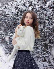 Russia, Moscow - 11/21/2020: Beautiful girl in a white sweater with a white cat in her arms in the winter in the forest.